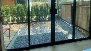 Ein Zimmer mit einer großen Schiebeglastür, die zu einem Außenbereich mit Kiesweg, Holzzaun und Blick auf ein Gebäude und Bäume führt.