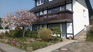 Un immeuble résidentiel de deux étages doté d’un balcon, d’un jardin avec des plantes en fleurs et d’un ciel dégagé.