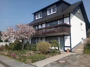 A two-story residential building with a balcony, a garden with flowering plants, and a clear sky. - Apartment Magnolie – with MeineCardPlus and New Furnishings for 4 Guests (Willingen (Upland))