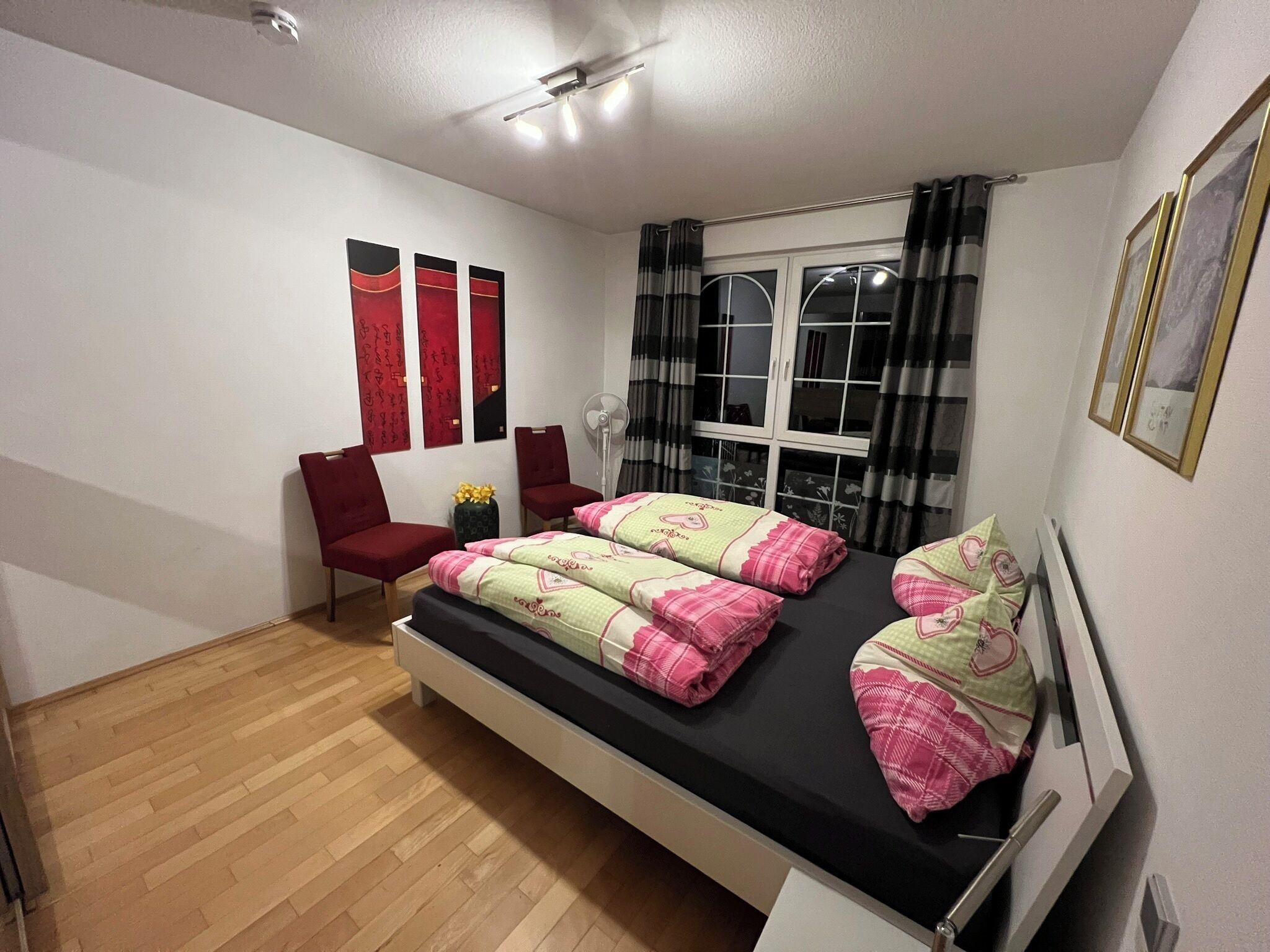 A bedroom with a bed, a chair, a fan, and a window with curtains.