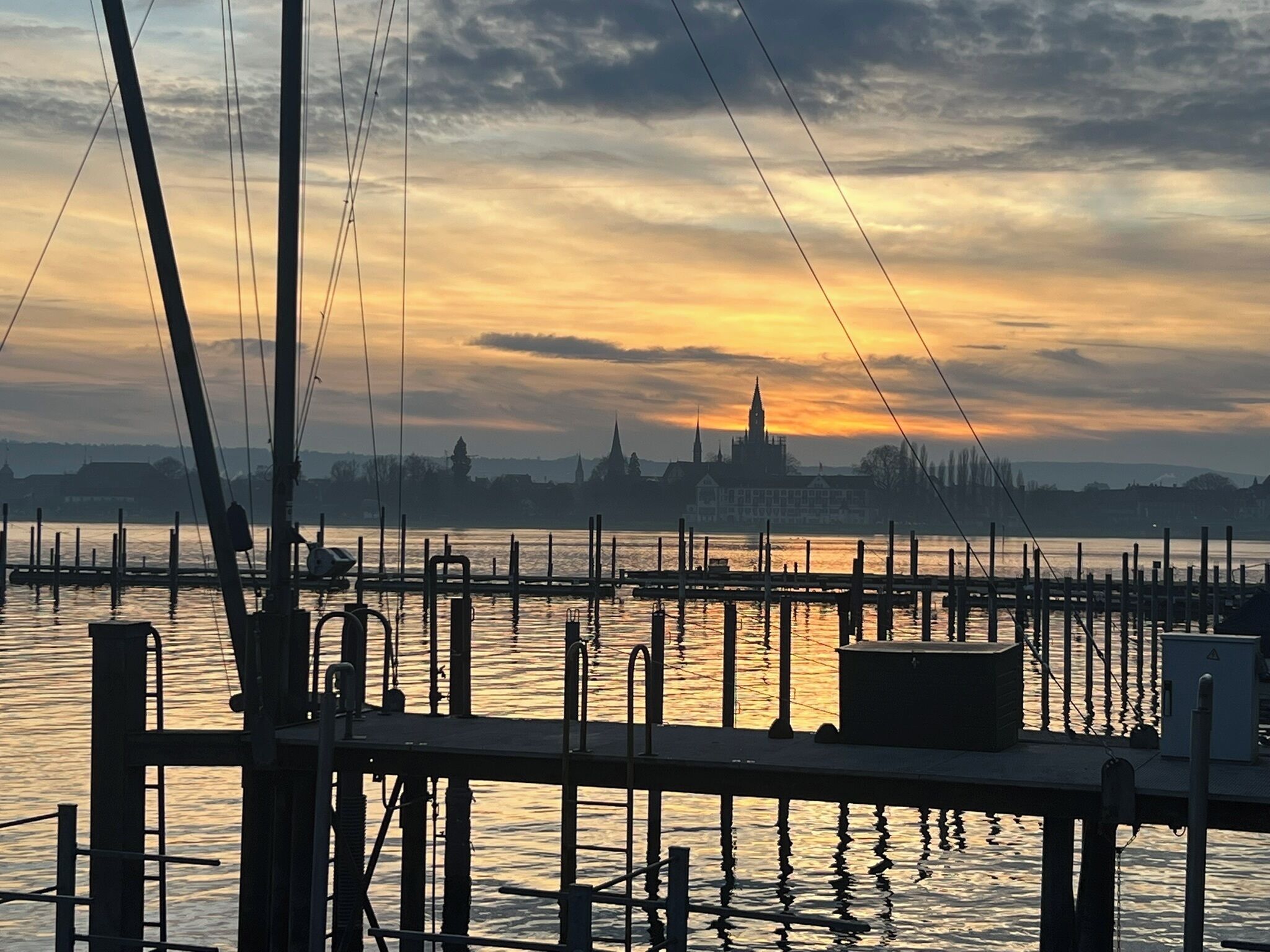 Blick auf das Wasser mit einer Anlegestelle, Booten und einer fernen Stadtlandschaft im Sonnenuntergang.