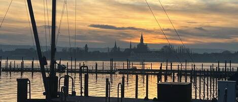 Blick auf das Wasser mit einer Anlegestelle, Booten und einer fernen Stadtlandschaft im Sonnenuntergang.