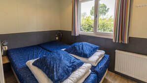 Una habitación con dos camas, una ventana con cortinas, un radiador y una mesita de noche pequeña con una lámpara.