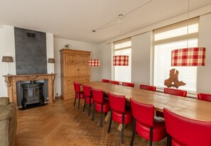 Een eethoek met een lange houten tafel, rode stoelen, een open haard en lampenkappen met ruitpatroon.