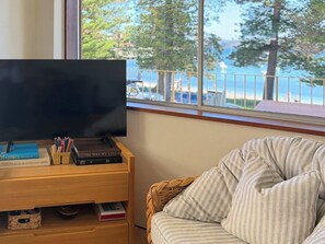 Ein Zimmer mit Fernseher auf einem Holztisch, einem Korbstuhl und Meerblick durch ein Fenster.