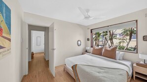 Ein Schlafzimmer mit einem Bett, einem Deckenventilator, einem Fenster mit Blick auf Palmen und einer Tür zu einem anderen Raum.
