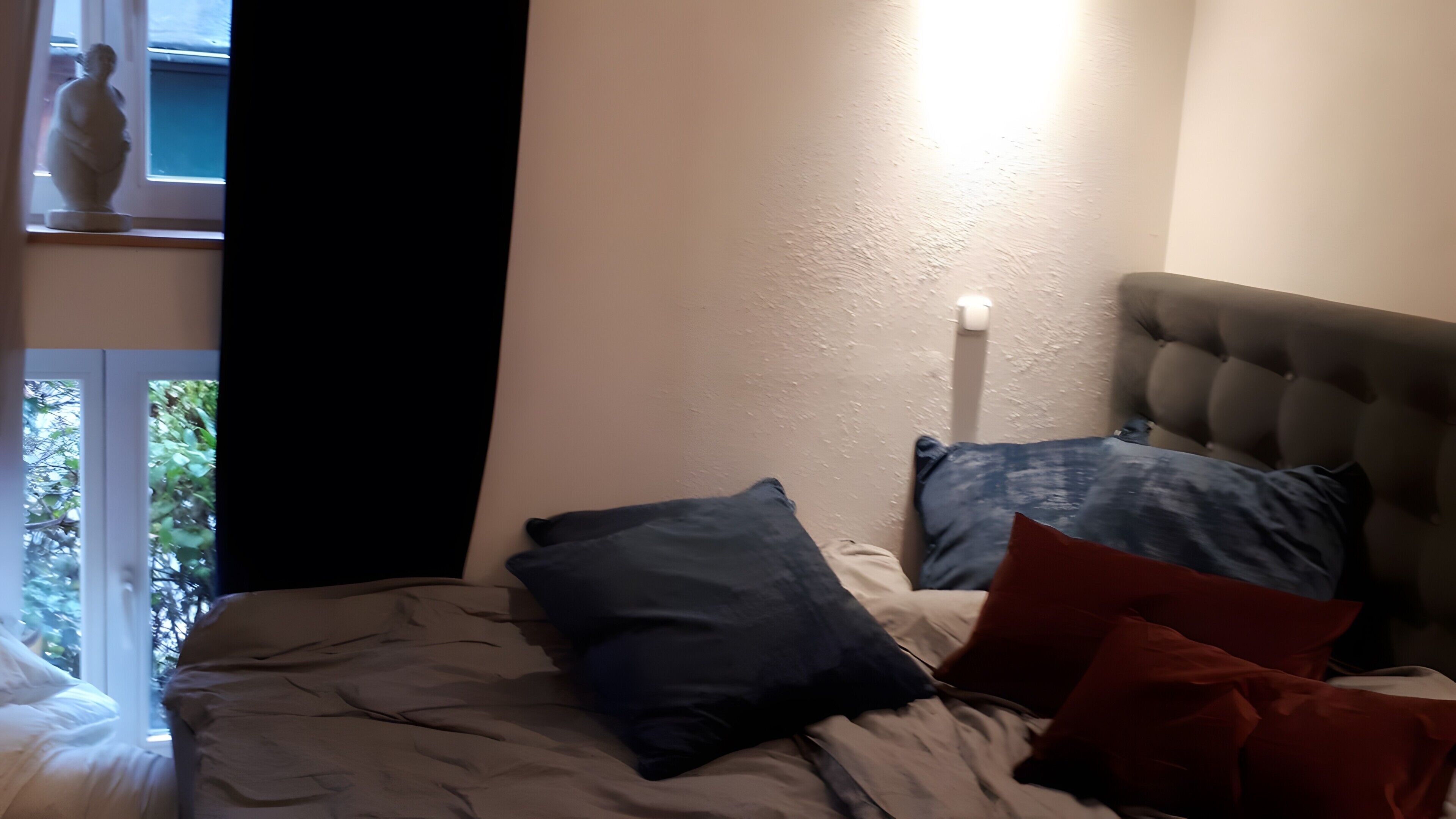 A bedroom with a bed, pillows, a window, and a wall-mounted light.