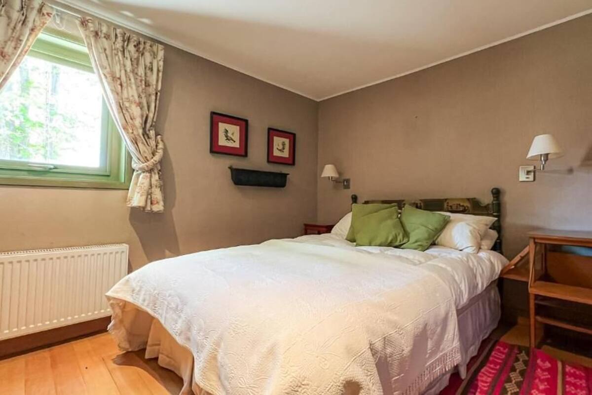 Ein Schlafzimmer mit einem Bett, einem fensterbelaubten Fenster, einem Heizkörper, einem Regal mit Bildern und einem Nachttisch mit Lampe.