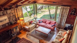 Un salon chaleureux avec une cheminée en pierre, un plafond en bois et de grandes fenêtres donnant sur un jardin.