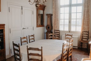 Une salle à manger avec une table ronde, des chaises en bois, un lustre, un miroir et une fenêtre avec des rideaux.