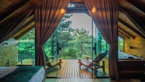 An attic bedroom with a wooden ceiling, a bed, two chairs, a window with curtains, and a view of trees.
