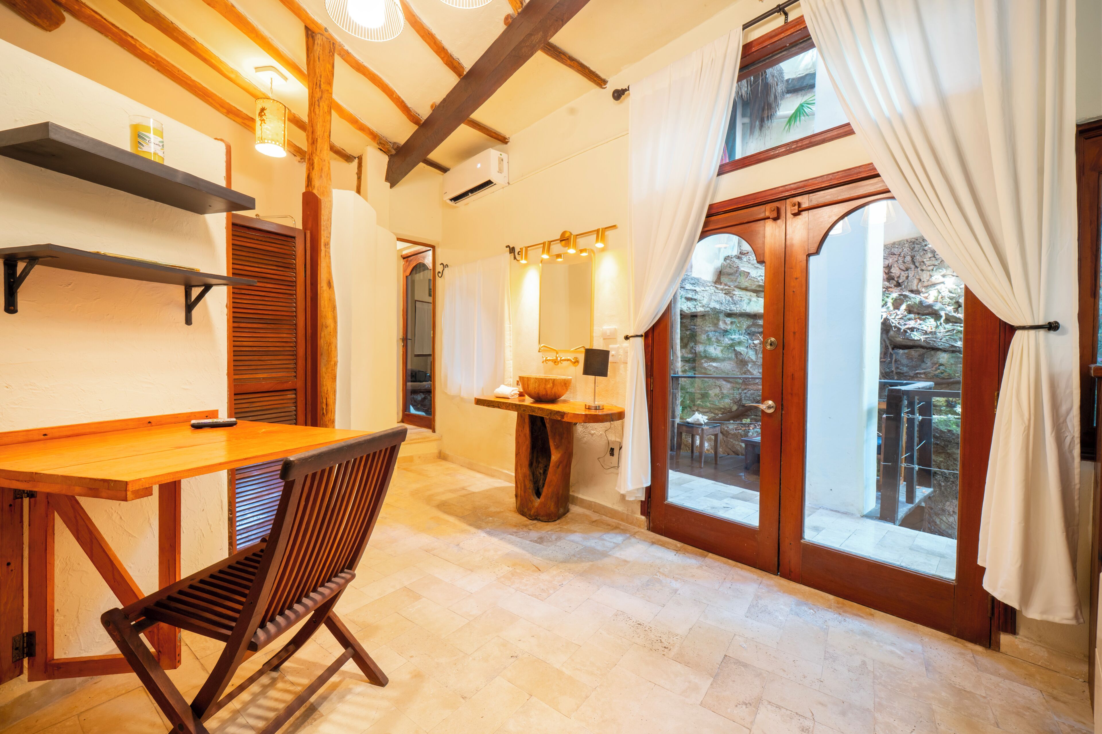 A room with a wooden table, a chair, a mirror, a window with curtains, and a stone wall outside.