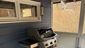 A portable Weber Spirit grill with a hose, positioned outside a house with a window and a wooden ceiling.