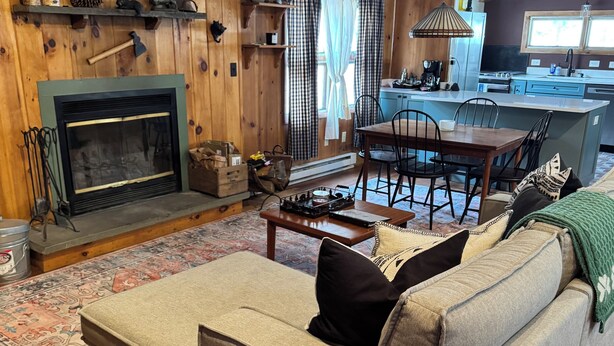 A cozy living room with a fireplace, wooden beams, a sofa, a dining table, and a kitchen area.