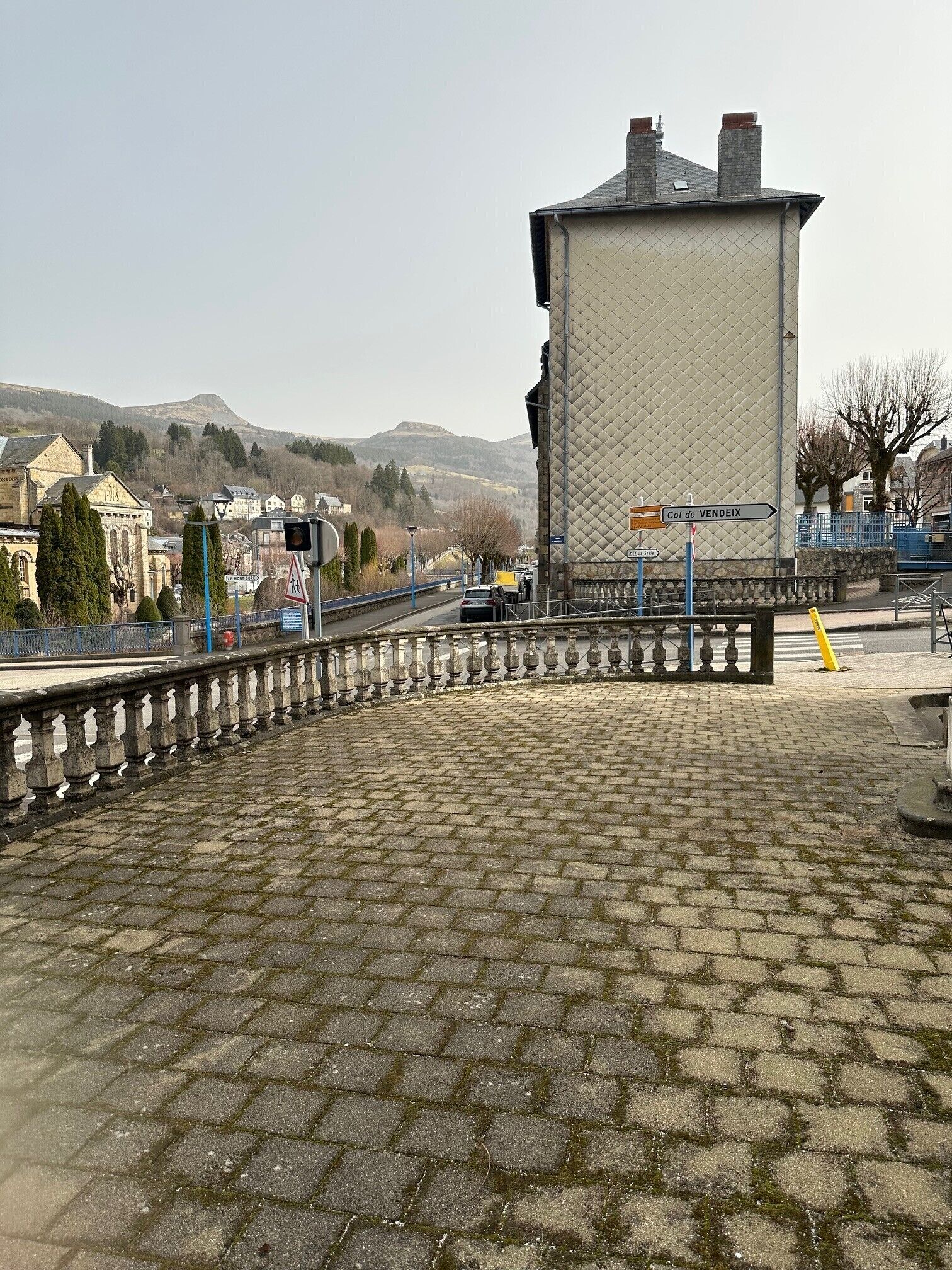 A cobblestone plaza with a stone balustrade, a tall building with a quilted facade, and a sign indicating direction to Col de Vendeix.