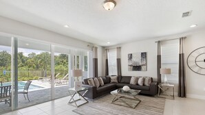 A spacious living room with a sectional sofa, a glass coffee table, and a view of a pool and greenery outside. - Last Minute Disney Getaway! Private Pool, Hot Tub & Game Room. Book Now! #15070 (Orlando)