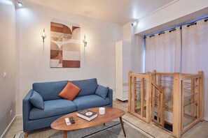 A modern living room with a blue sofa, a coffee table with a book and remote, a wall-mounted abstract art piece, and a wooden staircase. - Charming Duplex Center of Paris (Paris)