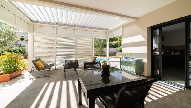 Ein überdachter Patio-Bereich mit einem Glastisch, zwei Stühlen, einem Topf mit Pflanze und Blick auf einen Swimmingpool.