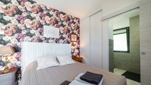 Ein Hotelzimmer mit blumigem Wandpapier, einem Bett mit Kissen und gefalteten Handtüchern, einem Nachttisch mit Lampe sowie einem Duschbereich.
