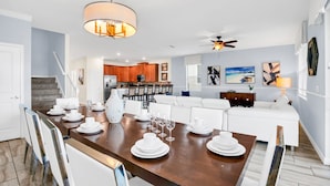 Un coin repas avec une table en bois dressée pour six personnes, des chaises blanches, un lustre, un ventilateur de plafond et une cuisine en arrière-plan.