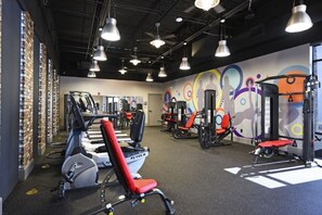 Ein Fitnessraum mit verschiedenen Trainingsgeräten, einer Wand mit farbenfrohen Kunstwerken und Pendelleuchten.