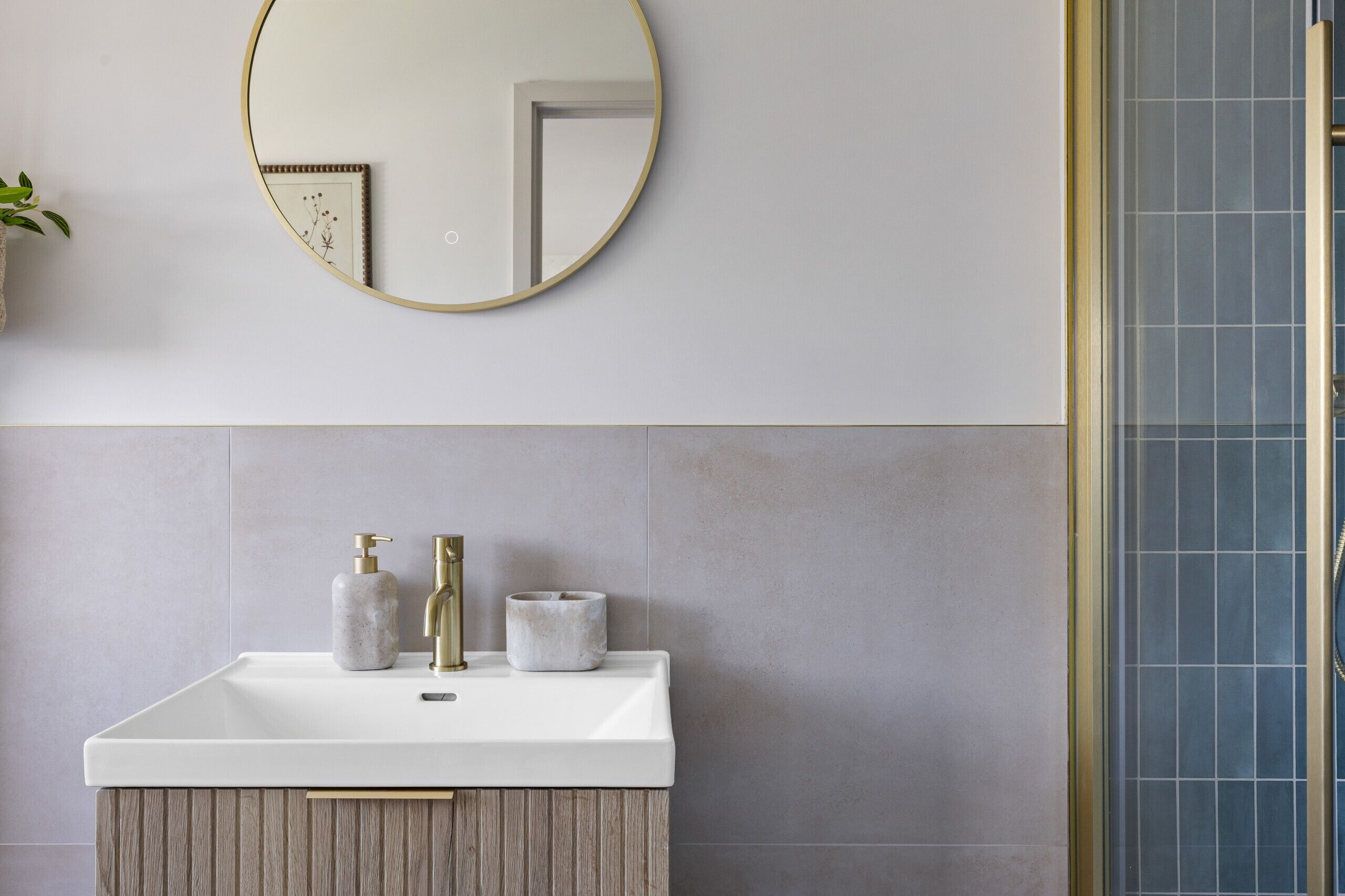 A bathroom with a round mirror, a white sink, a marble soap dispenser, a gold faucet, and a wooden vanity.