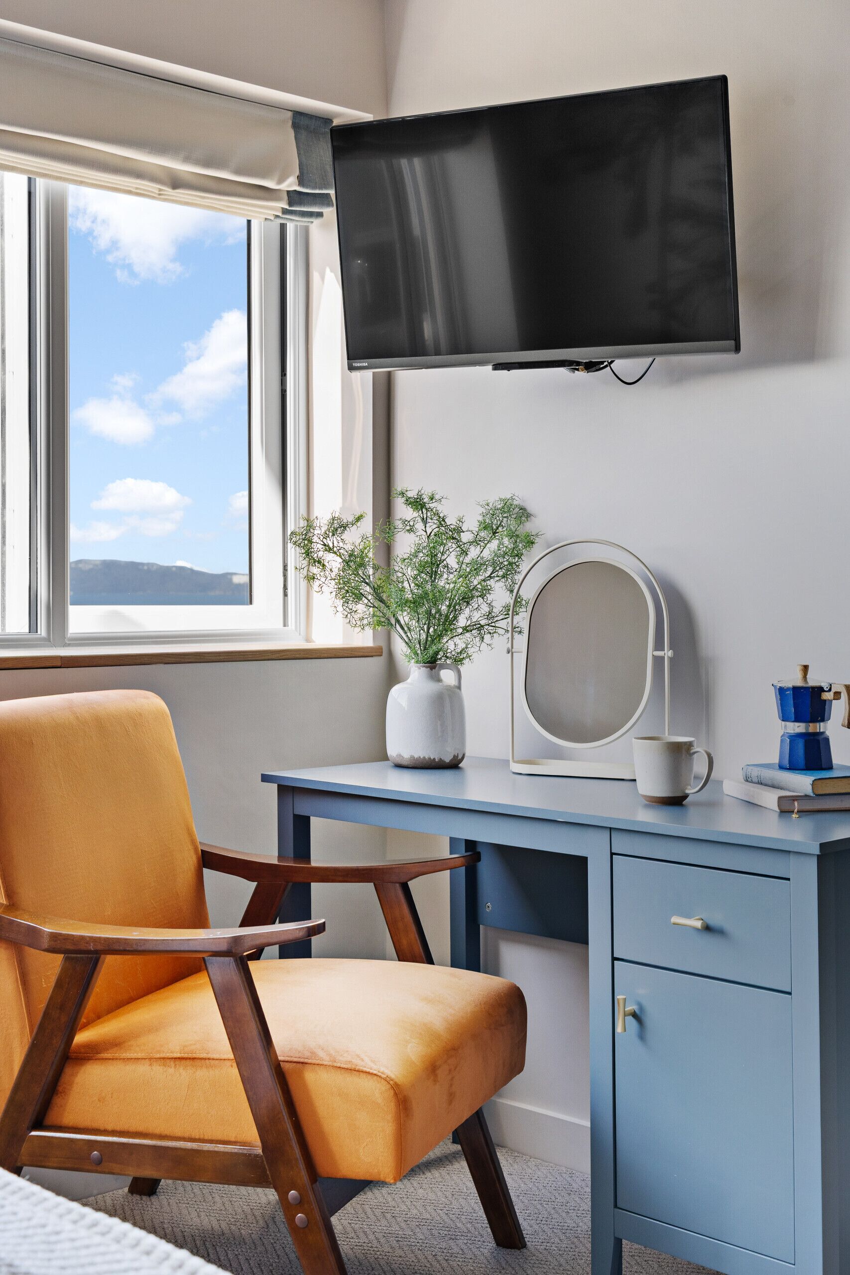 Een kamer met een aan de muur bevestigde televisie, een bureau met stoel, een klein kastje, een potplant en een raam met uitzicht.