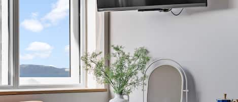 Een kamer met een aan de muur bevestigde televisie, een bureau met stoel, een klein kastje, een potplant en een raam met uitzicht.