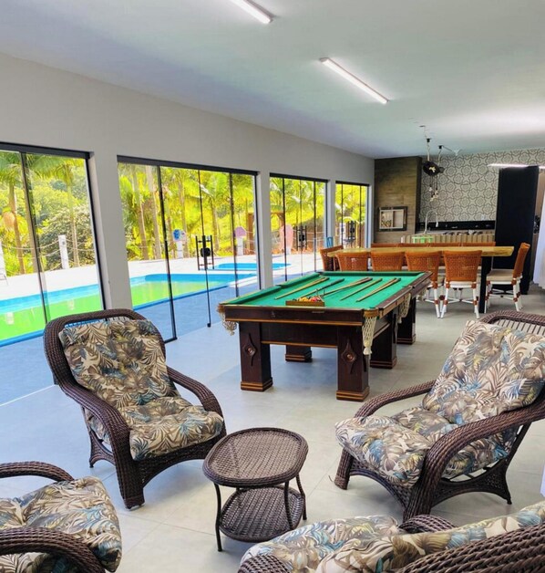 A pool table in a room with large windows, wicker chairs, and a dining area. - Sítio Moderno do Zizo com Jacuzzi (Angelina)