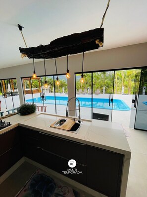 A modern kitchen with a large island, a built-in sink, and a view of a swimming pool. - Sítio Moderno do Zizo com Jacuzzi (Angelina)