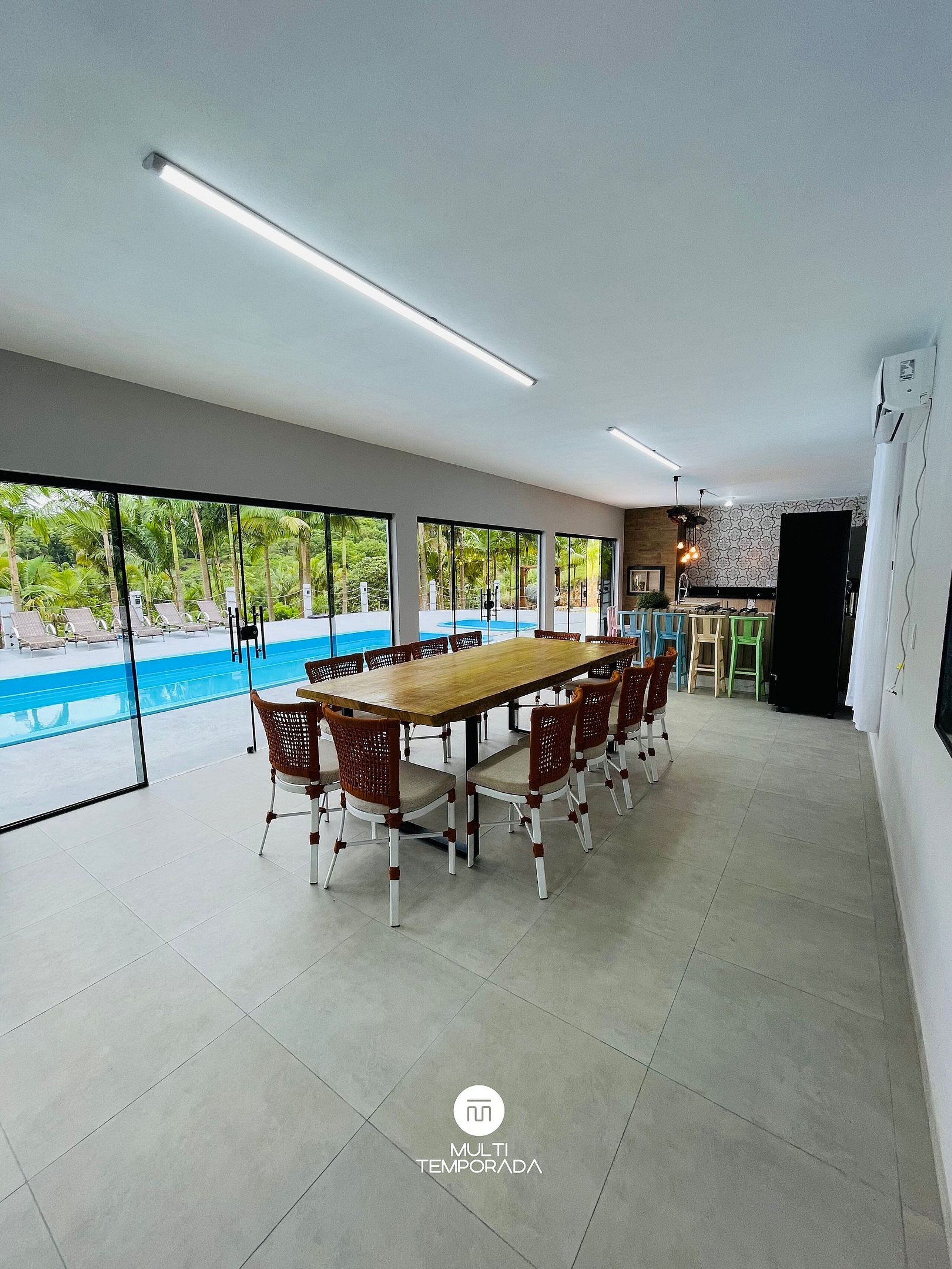 A spacious indoor area with a long wooden table surrounded by wicker chairs, large windows offering a view of a swimming pool and palm trees, and a kitchen area with modern appliances.