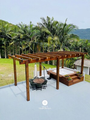 Een houten pergola met een jacuzzi en zitgedeelte, omringd door weelderige begroeiing en een glooiend landschap.