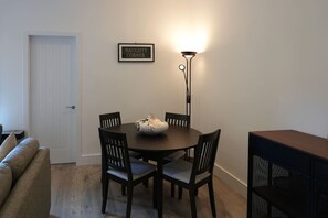 Un comedor con una mesa redonda, cuatro sillas, un jarrón, un cartel enmarcado, una lámpara de pie, un sofá y un mueble para televisor.