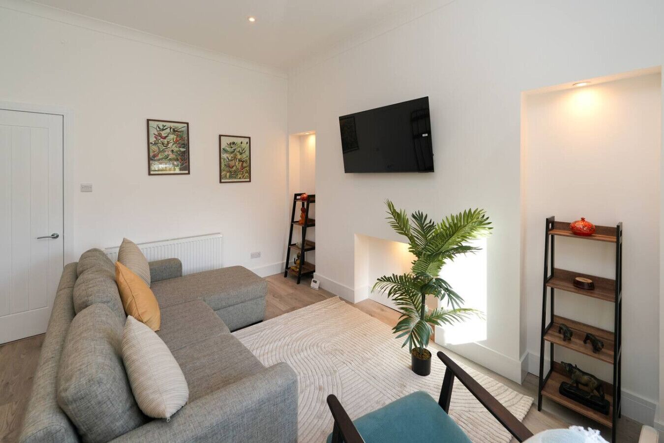 A living room with a sofa, a TV mounted on the wall, a plant, and a shelf with decorative items.