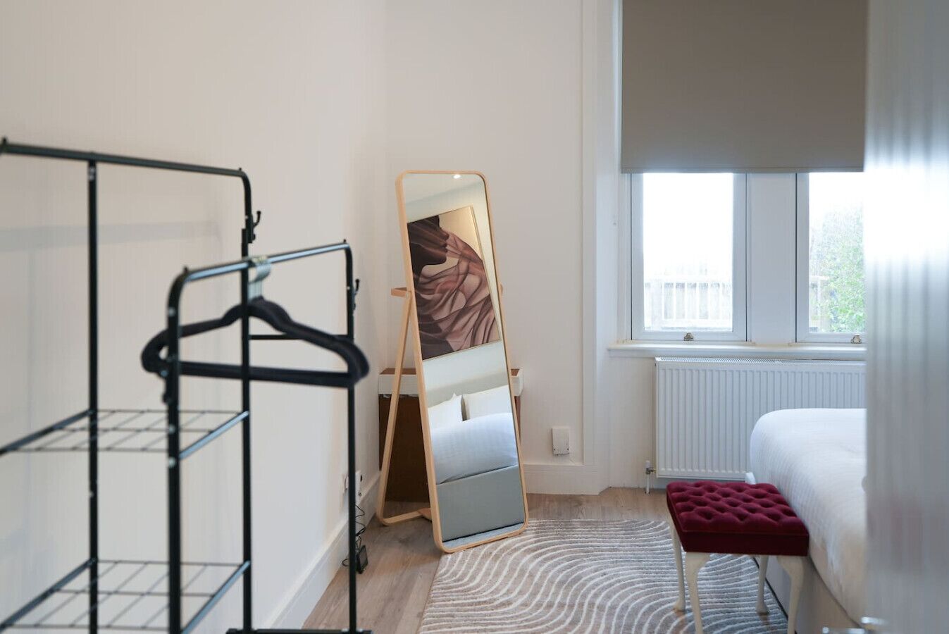 A hotel room with a bed, a red ottoman, a mirror, a radiator, and a window.