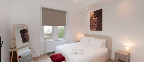 A bedroom with a large bed, a bedside table, a nightstand, a mirror, and a window with blinds.