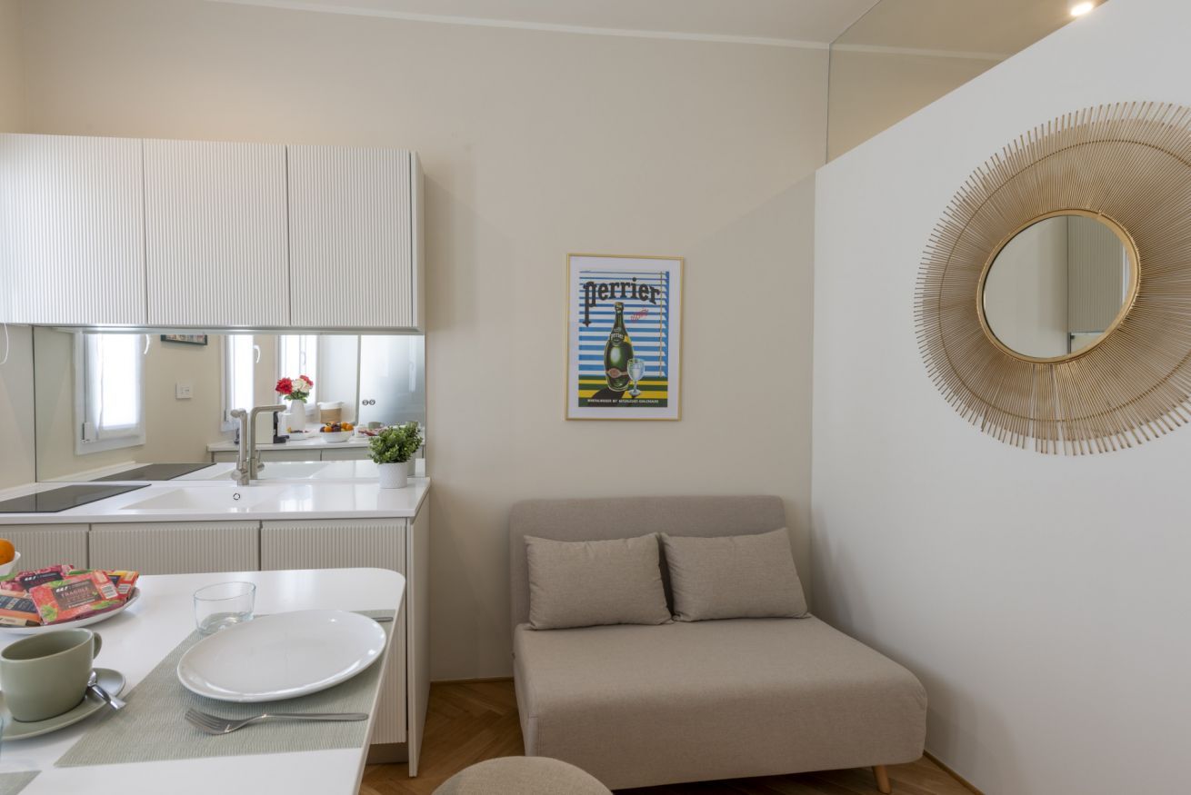 A compact living space with a kitchenette, a sofa, a dining table with a plate and utensils, a mirror, and a poster on the wall.