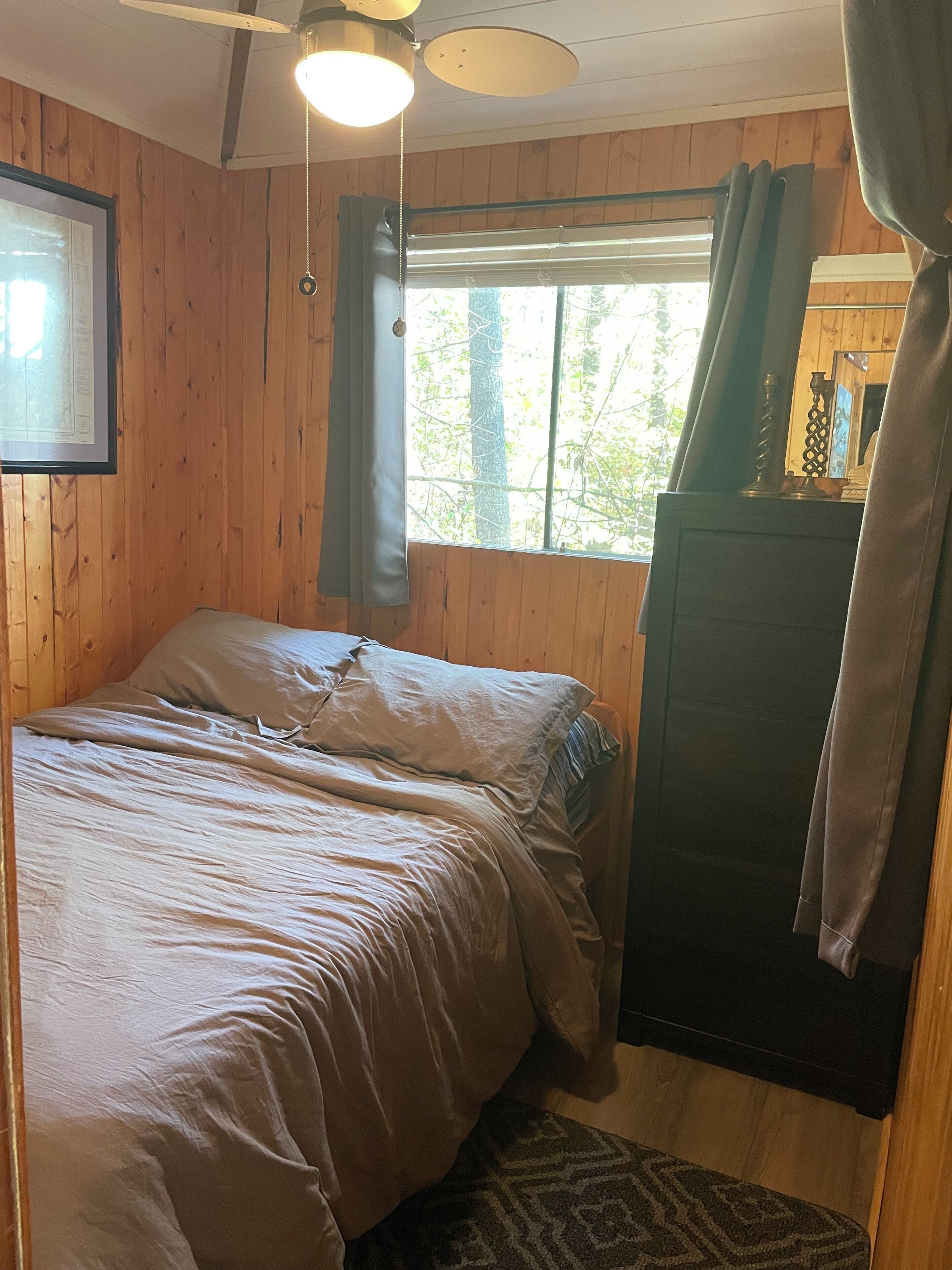 Una stanza con rivestimento in legno, un letto, una finestra con tende, una ventola a soffitto e un quadro appeso al muro.