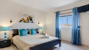 A bedroom with a bed, bedside lamps, a painting, a window with curtains, and a television on the wall. - Apartment 'Algarve House' with Sea View, Wi-Fi and Air Conditioning (Albufeira)