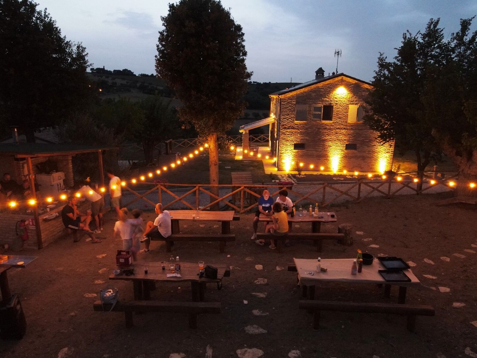 Un'area pranzo all'aperto con tavoli da picnic e luci decorative appese, circondata da alberi e un edificio in pietra sullo sfondo.