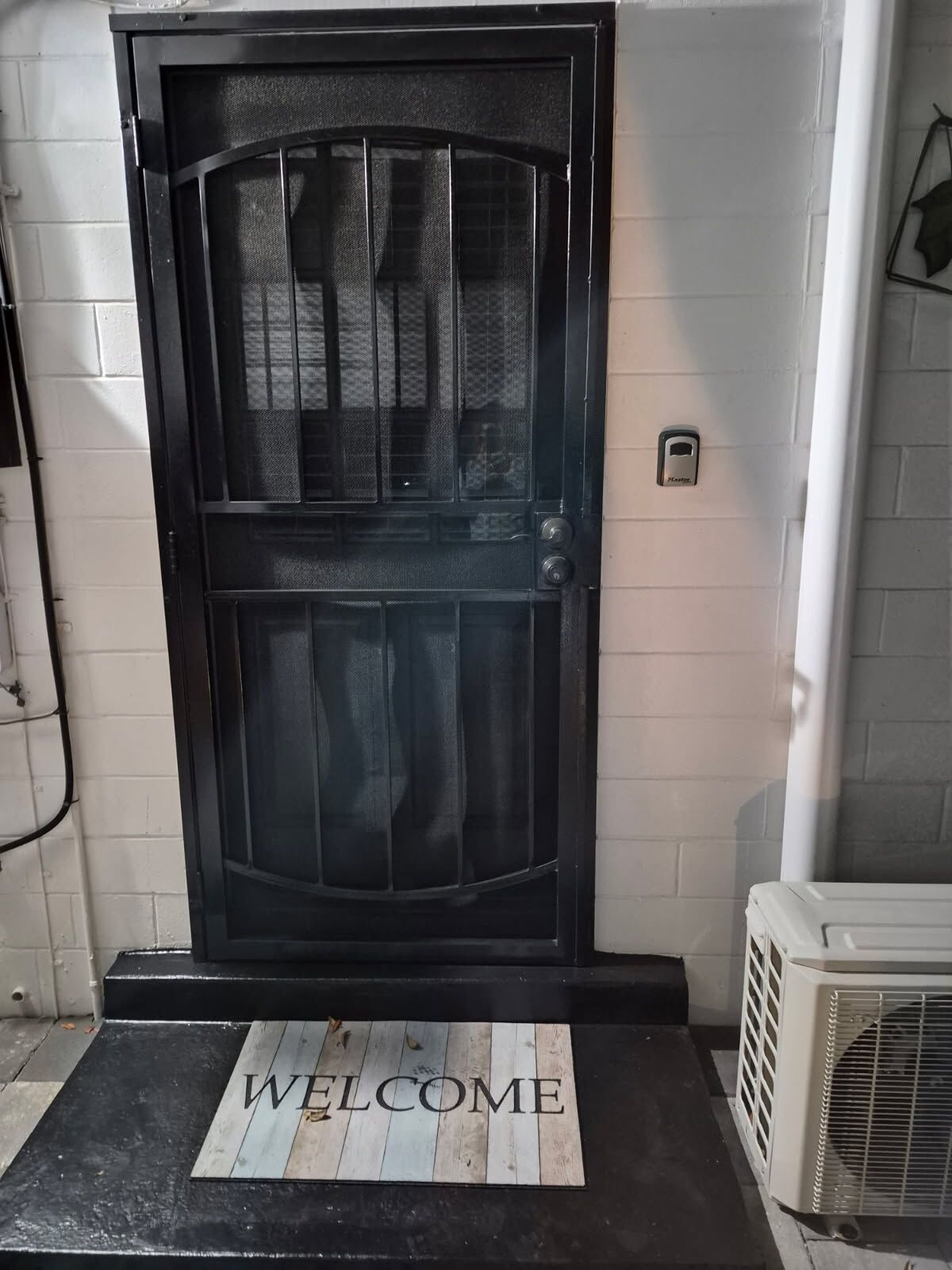 A black door with a mesh screen, a 'WELCOME' mat, and a white air conditioning unit.