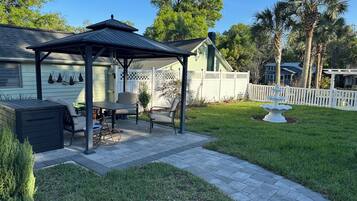 Un patio techado con una mesa y sillas, un pequeño jardín con una fuente blanca y una cerca de piquetes blancos.