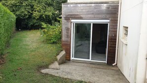A residential property with a sliding glass door entrance, a concrete pathway leading to the door, and a grassy yard with bushes and trees. - Self-contained studio at the edge of Ecouves (Cuissai)