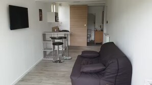 A modern living room with a dark sofa, a small kitchen area with a bar stool, and a TV mounted on the wall. - Self-contained studio at the edge of Ecouves (Cuissai)