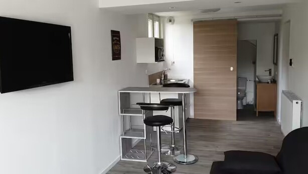 A modern kitchen with a bar area, a flat-screen TV mounted on the wall, and a wooden door leading to another room. - Self-contained studio at the edge of Ecouves (Cuissai)