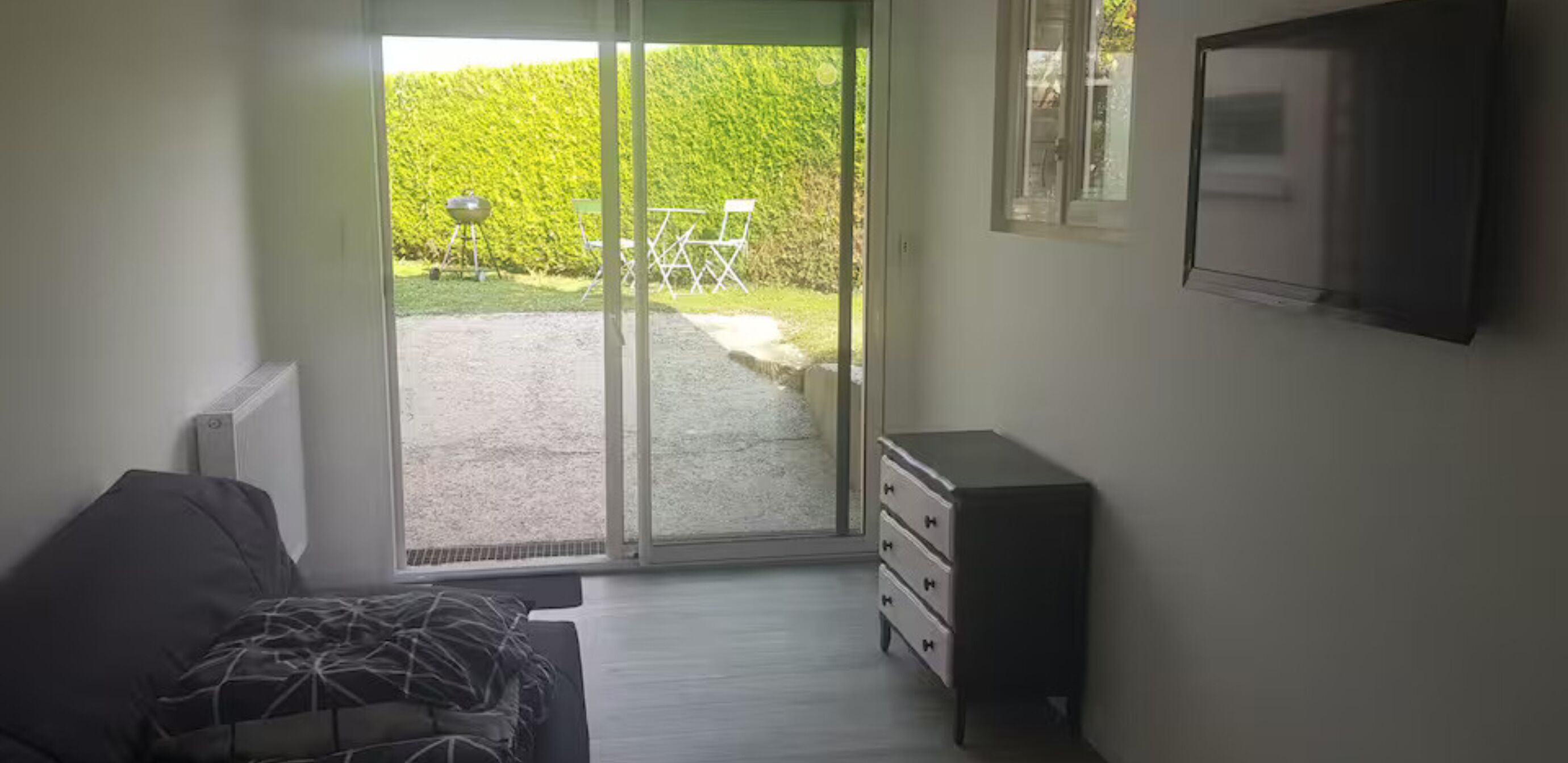 A room with a bed, a dresser, a sliding glass door leading to an outdoor area with a barbecue grill and chairs.