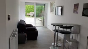 A modern living room with a sofa, a small table with chairs, a radiator, and a sliding glass door leading to an outdoor area. - Self-contained studio at the edge of Ecouves (Cuissai)