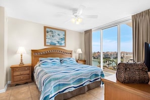 A hotel room with a large bed, two bedside tables with lamps, a ceiling fan, a large window with a view, and a wicker trunk. - NEW! Terrace at Pelican Beach 401 (Destin)