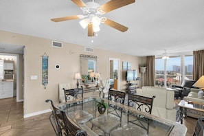 A spacious living room with a glass dining table, a ceiling fan, and a view of the city. - NEW! Terrace at Pelican Beach 401 (Destin)