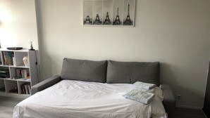 Un dormitorio con cama, cabecera gris, ropa de cama blanca, una estantería y un cuadro colgado en la pared de la Torre Eiffel.